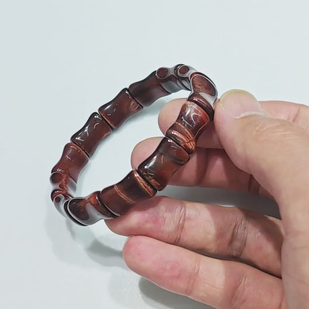 Hand holding natural red tiger eye elastic bracelet with bamboo-shaped bead units (15mm length), deep red-brown base and golden-red bands — fits standard 18cm wrist, unisex design