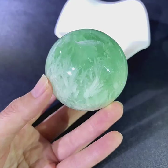 Hand holding natural green fluorite sphere with feather inclusions, demonstrating size, weight, and tactile texture