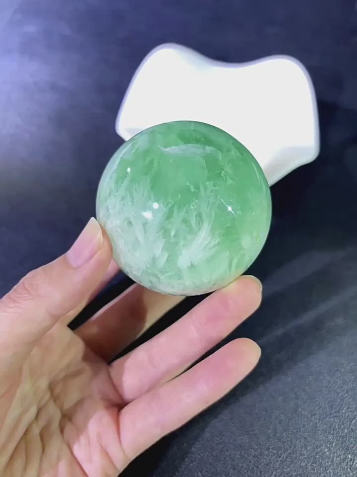Hand holding natural green fluorite sphere with feather inclusions, demonstrating size, weight, and tactile texture