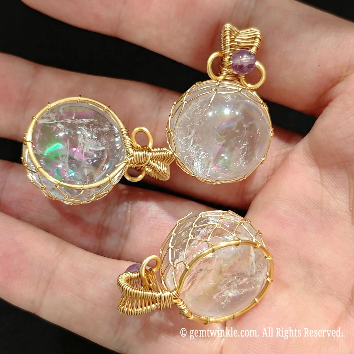 Pair of gold wire wrapped clear quartz sphere pendants resting on fingers showcasing internal rainbow reflections