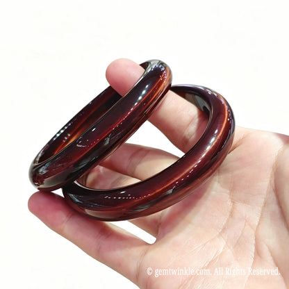Pair of solid red tiger eye bangles held in hand showcasing deep mahogany red luster and silky reflections