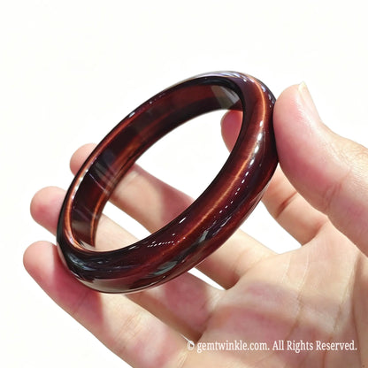 Angled hand view of a solid red tiger eye gemstone bangle showing smooth inner contour and polished finish