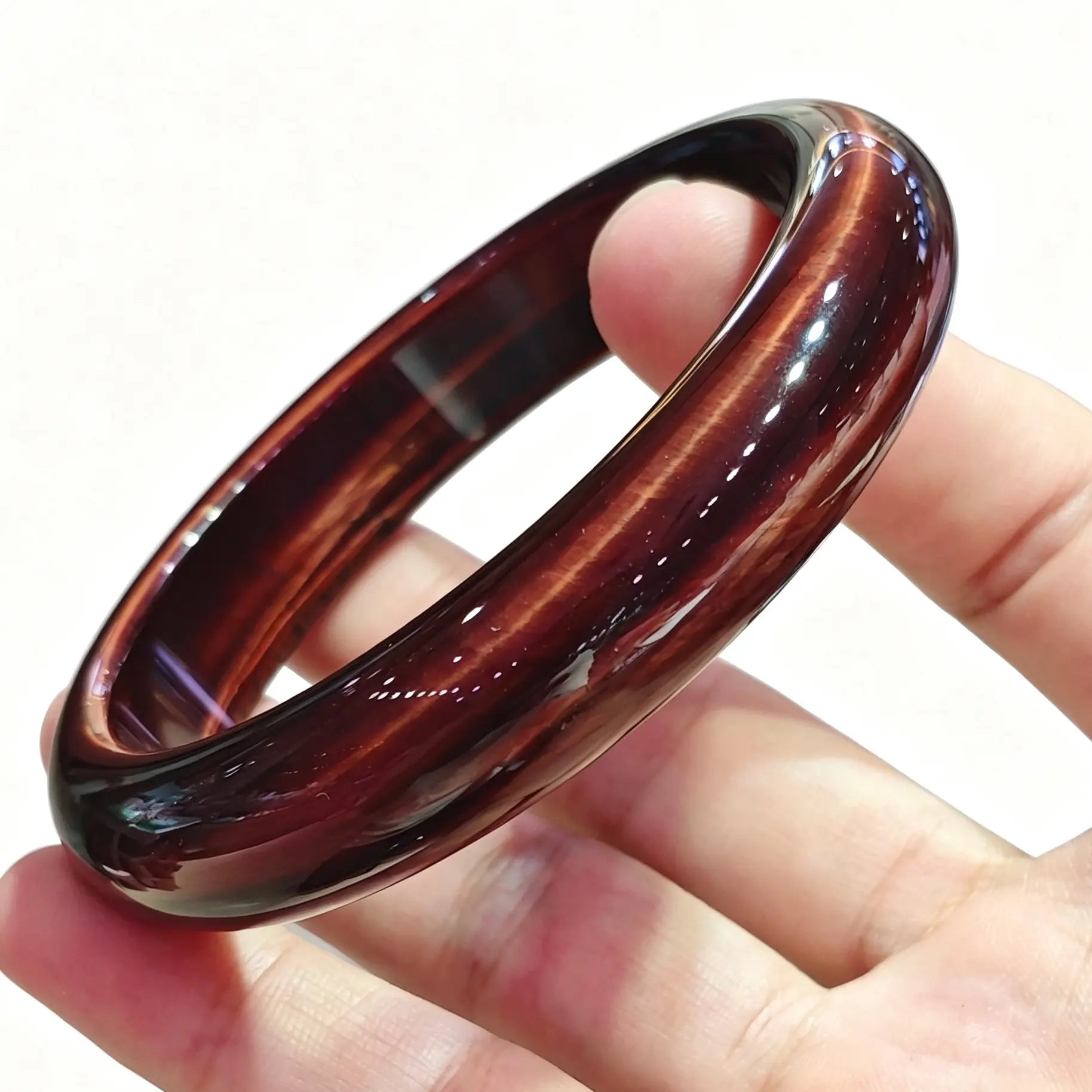 Fingers holding a smooth solid red tiger eye bangle displaying rich crimson mahogany color and silky chatoyancy