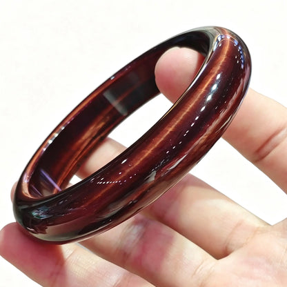 Fingers holding a smooth solid red tiger eye bangle displaying rich crimson mahogany color and silky chatoyancy