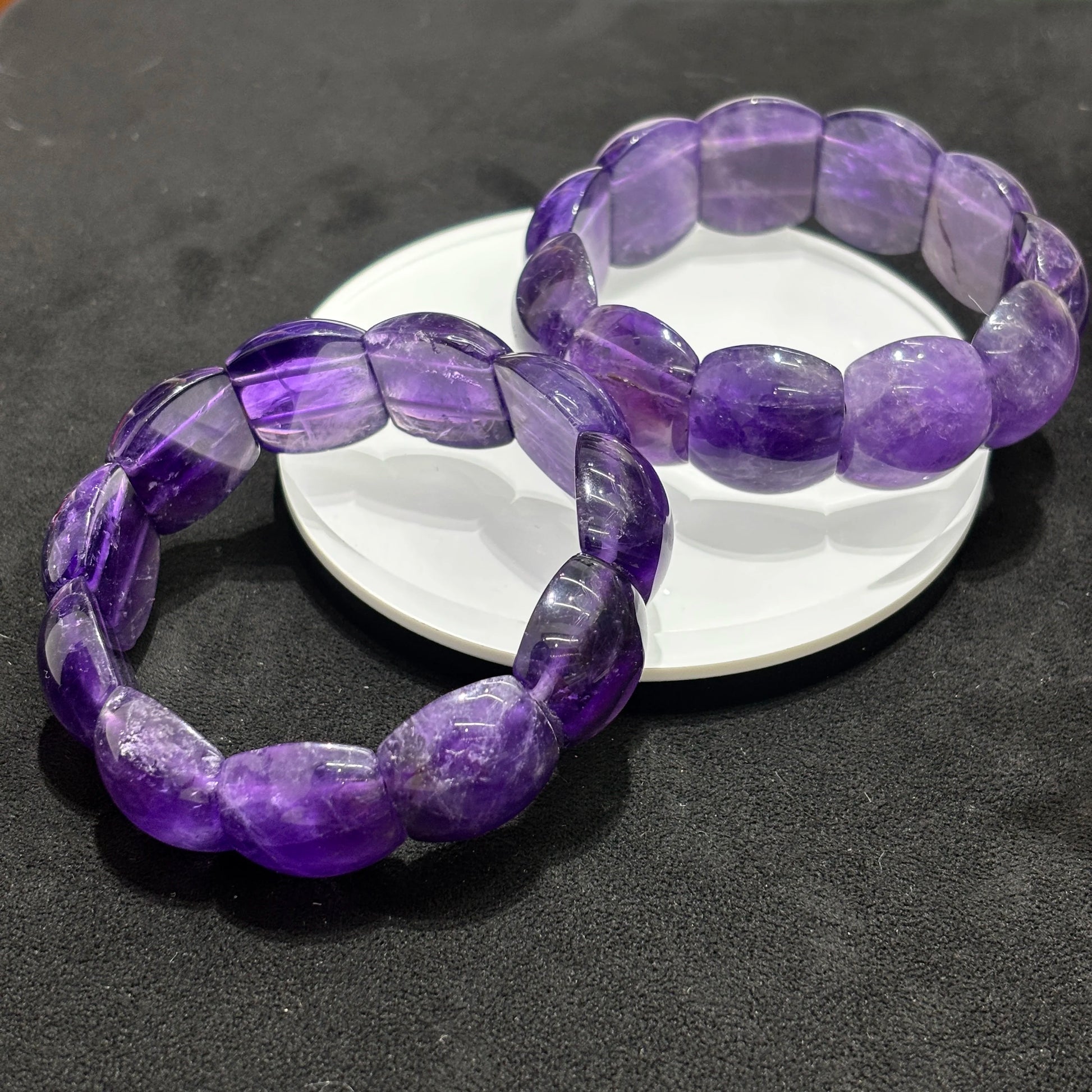 Side view of two natural amethyst rectangle bead bangle bracelets on a white dish, displaying the thickness and faceted edges of the beads.