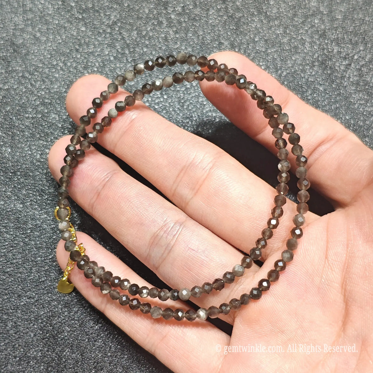 Hand holding 3mm micro faceted Silver Sheen Obsidian necklace strand showing dark charcoal glass with silver luster