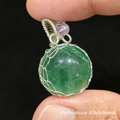 Front view of silver wire wrapped green fluorite sphere pendant held in hand showing intricate net structure