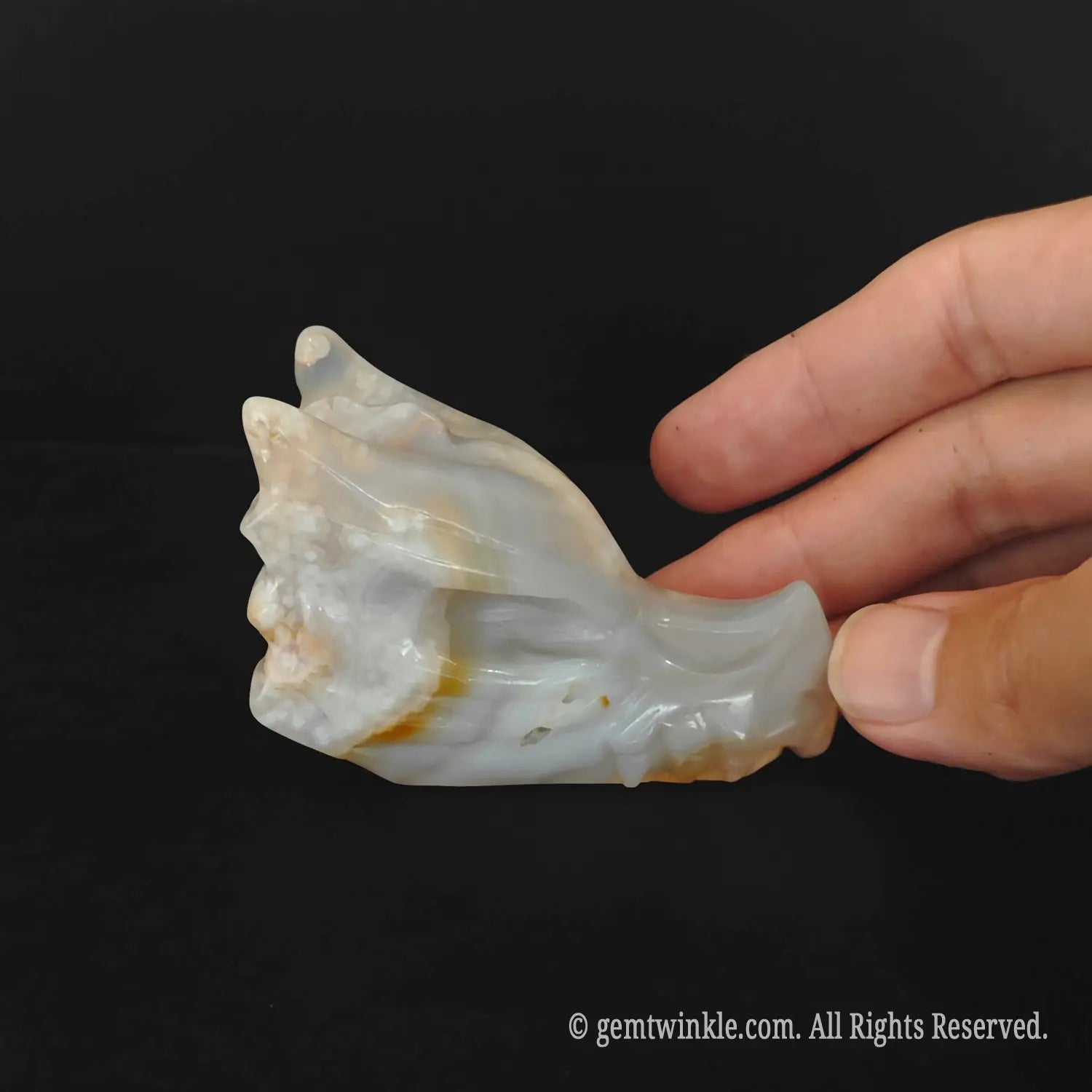 A side profile view of a 6cm flower agate dragon skull figurine held gently by a hand to demonstrate its pocket size and horn details