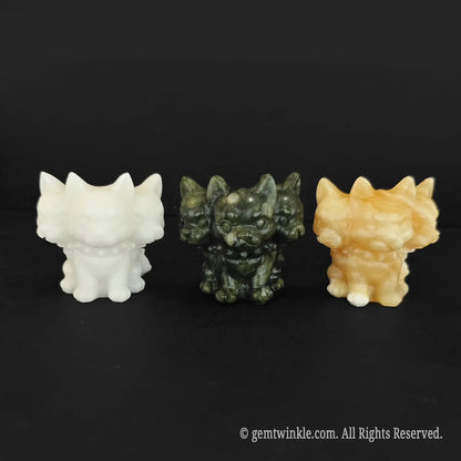 Three chibi three headed dog Cerberus figurines lined up horizontally on a black surface showing white marble green serpentine and yellow calcite variants