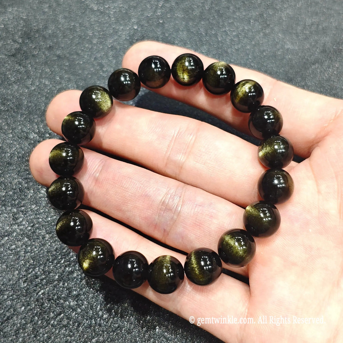 Full ring view of a premium select gold sheen obsidian bracelet resting on hand over textured grey fabric